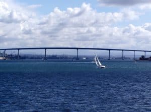 A white sailboat glides on dark blue water with a long bridge and cloudy sky in the background.